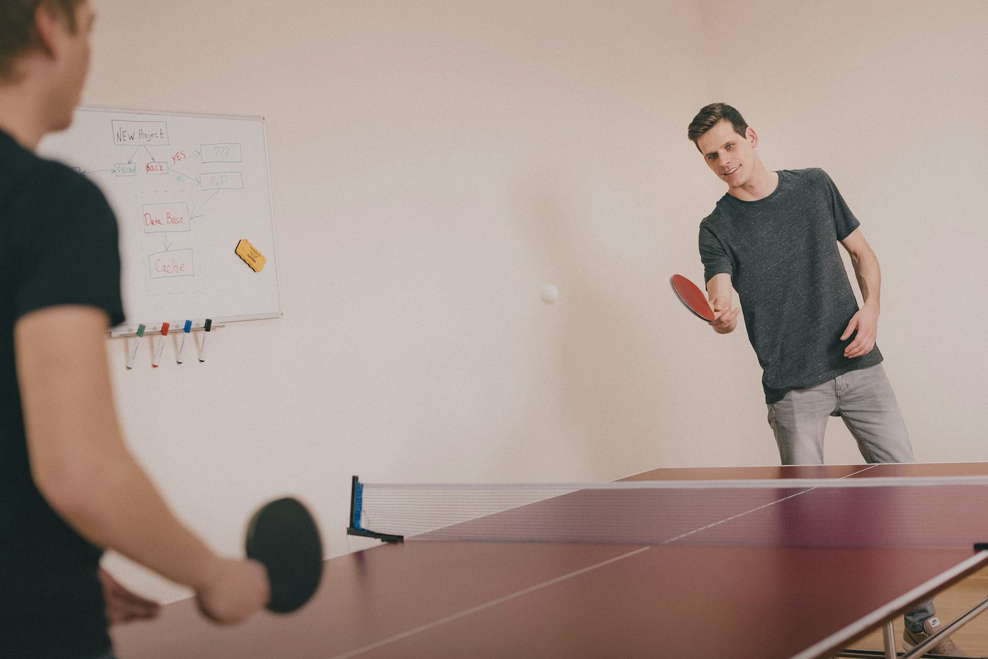 Two men play table tennis in an office with a whiteboard behind them.