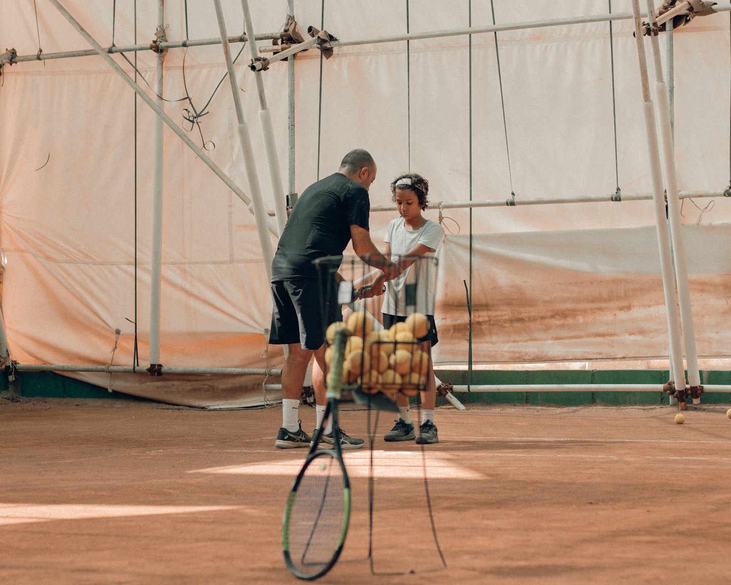 Tennis coach instructs a young player on an indoor clay court.