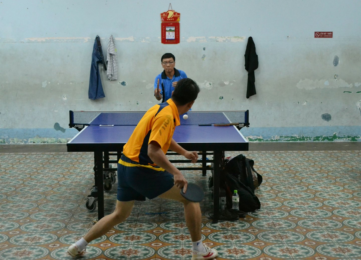 Two men playing table tennis indoors, one about to hit the ball.