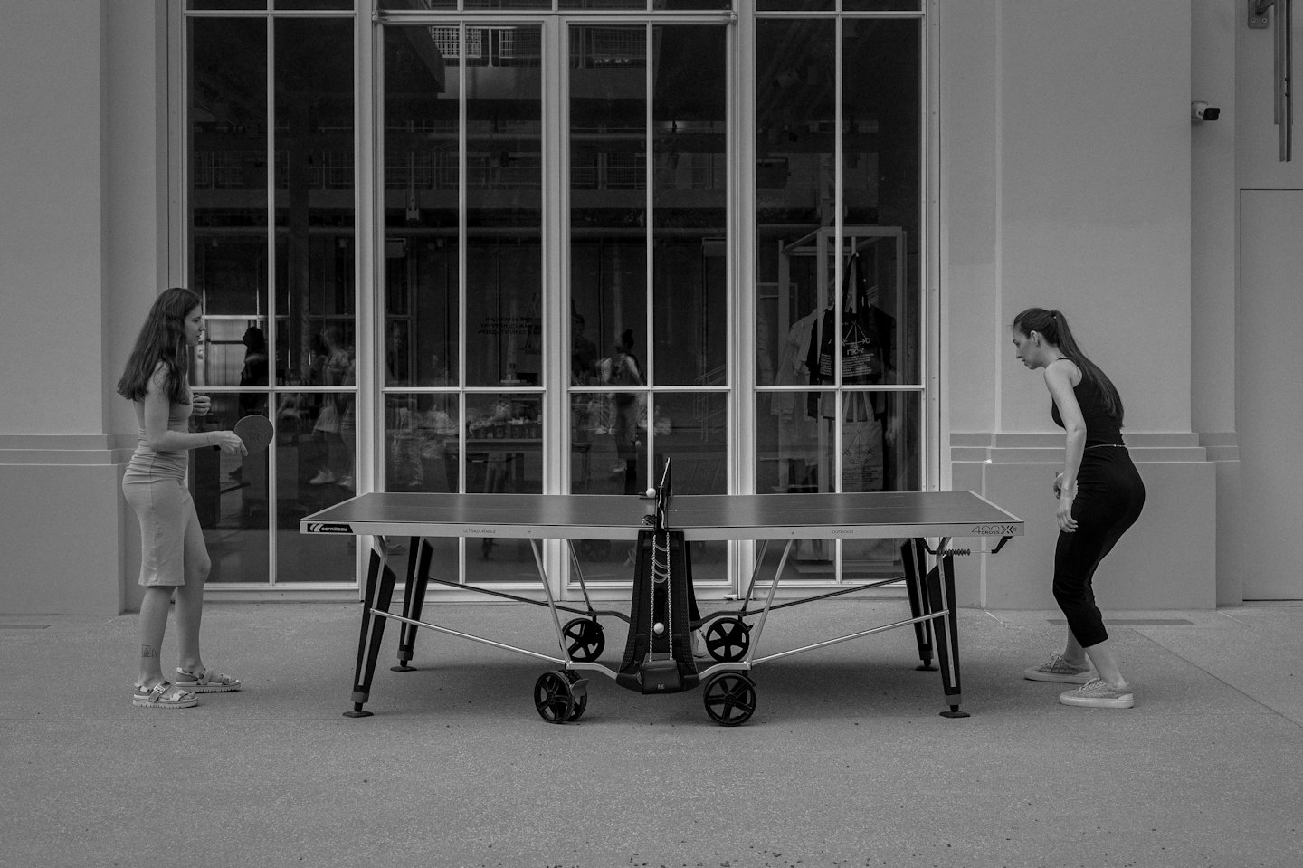 Two women playing table tennis outdoors with a building behind them.